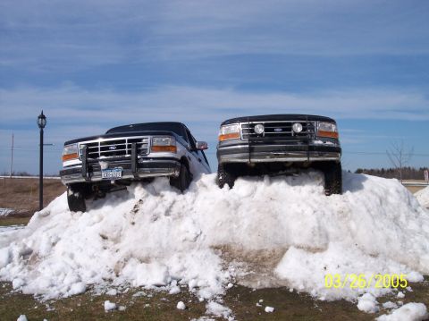 1995 Ford Bronco - Two 1995 Ford Broncos are parked atop a large snow pile under a bright blue sky.