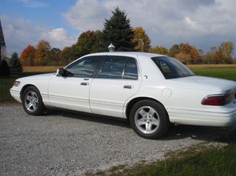 1996 Mercury Grand Marquis - A white sedan is parked outdoors on a gravel surface with trees and a cloudy sky visible.