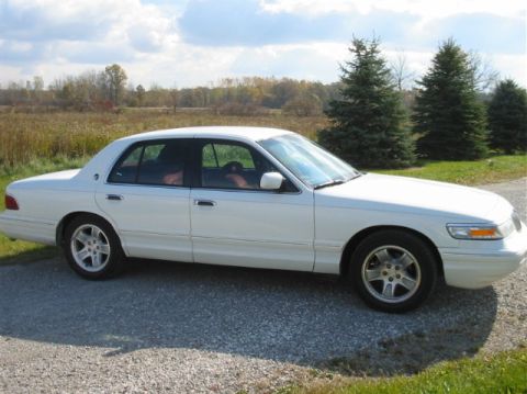 1996 Mercury Grand Marquis - A white 1996 Mercury Grand Marquis sedan is parked on a gravel surface with trees and fields in the background.