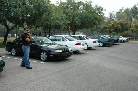 1991 Ford Taurus - A line of sedans are parked in a lot, with a man standing next to the first car on his phone.