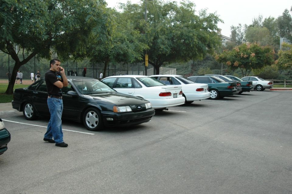 Man on phone next to black 1991 Ford Taurus, parked in a line with other sedans