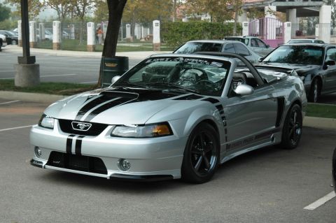 1991 Ford Taurus - A silver convertible Ford Mustang with black racing stripes is parked in a lot with other vehicles.
