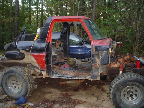 1981 Ford Bronco - Side view of a stripped red-and-black Ford project 4x4 in the woods, with its doors and interior removed to expose a bare cab and rusty flo…