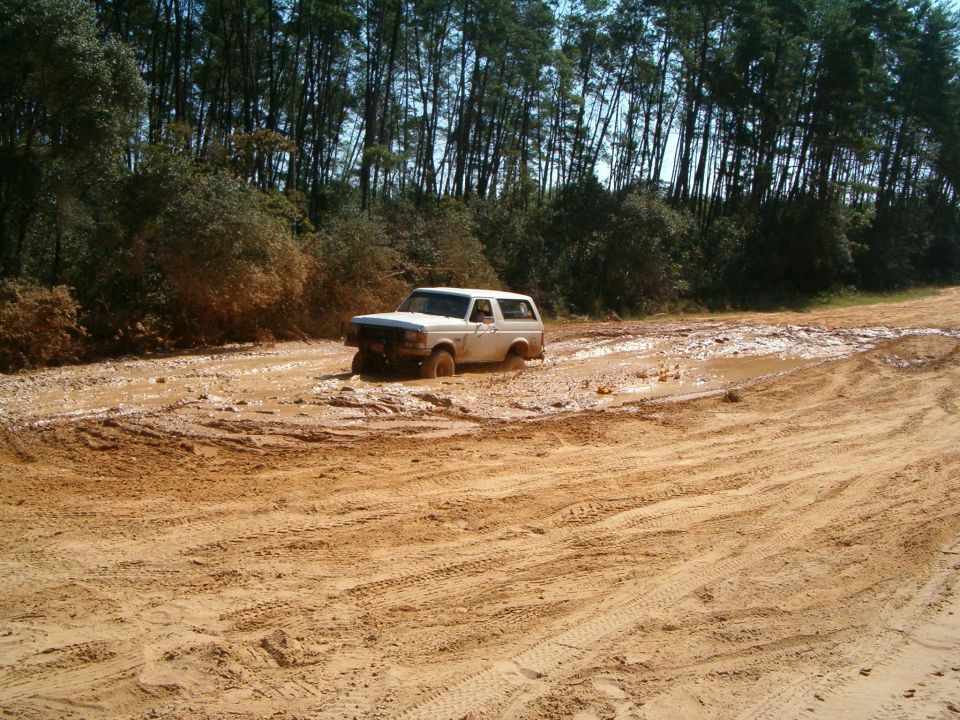 White Ford Bronco driving through a muddy trail with trees in the background