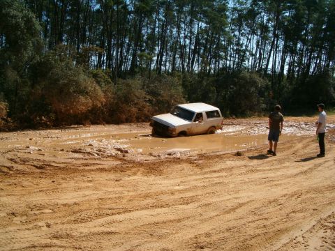 1991 Ford Bronco - A white SUV is stuck in a deep mud puddle on a dirt road, with two onlookers observing the situation.