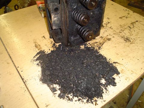 1981 Ford Bronco - Engine cylinder head removed from a vehicle, with a pile of dark, granular debris on the workbench.