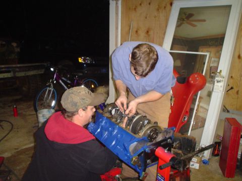 1981 Ford Bronco - Two individuals are assembling an engine block on a stand in a dimly lit garage.