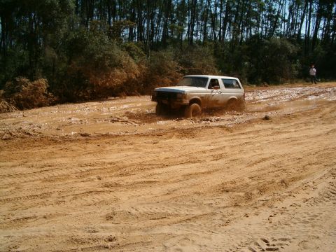 1991 Ford Bronco - A white 1991 Ford Bronco powers through a challenging muddy terrain, kicking up dirt and water.