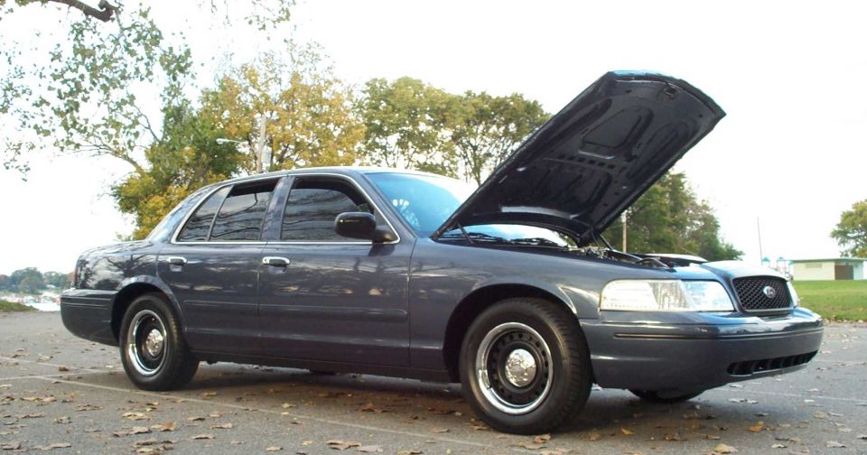 Dark blue-gray Ford Crown Victoria sedan with hood open, front three-quarter view in a lot