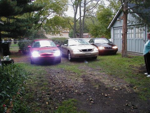 1997 Ford Taurus - Three sedans are parked in a grassy driveway next to a garage, with a woman standing to the right.