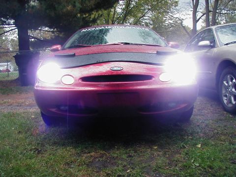 1997 Ford Taurus - A red 1997 Ford Taurus is shown from the front with its headlights illuminated.