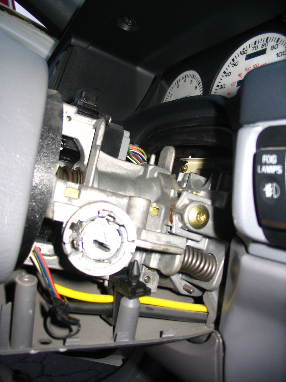 Car's ignition cylinder and surrounding mechanical parts, with dashboard gauges visible