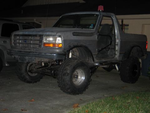 1992 Ford Bronco - Night three-quarter front shot of a heavily lifted gray Ford Bronco with the doors and rear top removed, sitting on tall military-style bia…