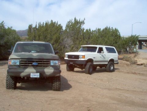 1988 Ford Bronco - Two Ford Broncos are parked on a dirt road with trees and a bridge in the background.