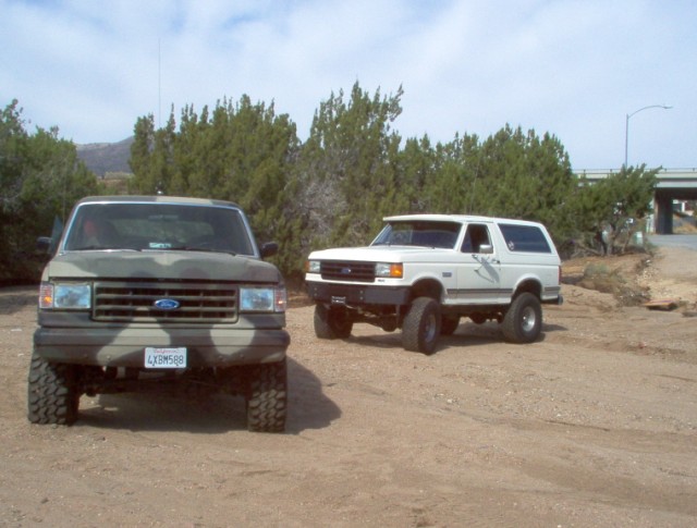 Two Ford Broncos, one in camouflage, one white, parked on dirt road