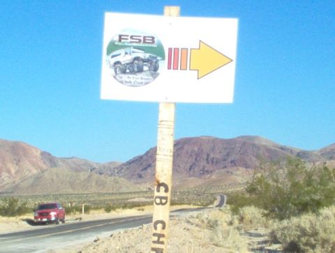 1988 Ford Bronco - A sign with an off-road club logo and directional arrow stands by a desert road, with a red SUV in the distance.