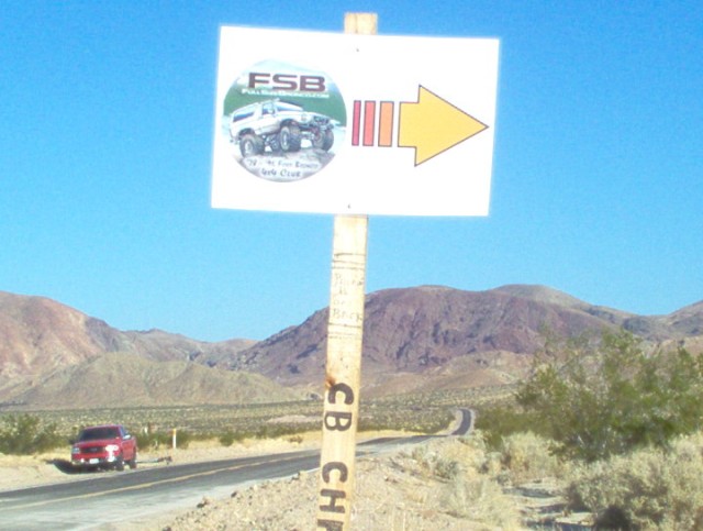 Sign with FSB logo showing a lifted SUV and a yellow arrow pointing right. A red SUV drives on a road in the desert