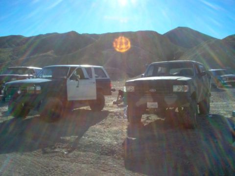 1988 Ford Bronco - Two Ford Broncos parked on dirt with mountains in background. One has driver door open.