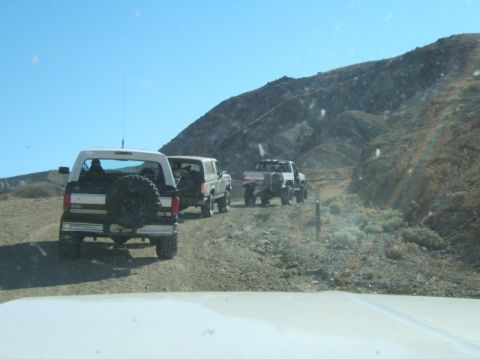 1988 Ford Bronco - Three SUVs driving on a dirt road in a desert landscape under a clear blue sky.