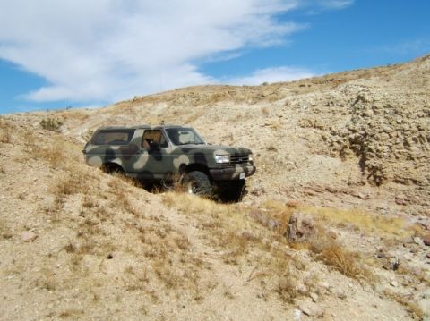 1988 Ford Bronco - A camouflaged SUV navigates a , arid terrain with sparse vegetation and layered rock formations.