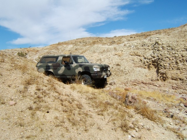 Camouflaged SUV driving up a rocky, dry hill under a blue sky with scattered clouds