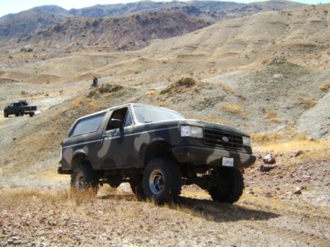 1988 Ford Bronco - A camouflaged Ford Bronco with lifted suspension navigates a dusty, arid landscape, with another vehicle visible in the background.