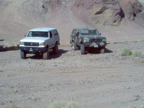 1988 Ford Bronco - Two SUVs, one white and one camouflaged, are parked off-road in a desert landscape.