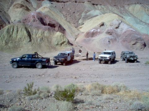 1988 Ford Bronco - Several off-road vehicles are stopped in a dusty, arid canyon with layered, colorful rock formations.