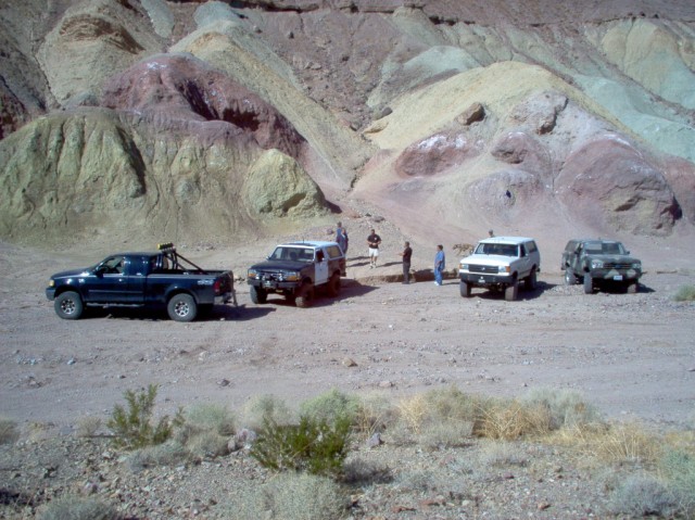Four off-road vehicles parked in a desert canyon with colorful hills in the background