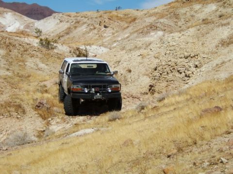 1988 Ford Bronco - A black and white Ford Bronco navigates a , dry, hilly terrain under a clear blue sky.