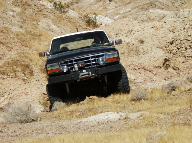 Black Ford Bronco with white top driving on a rocky, dry hillside. Aftermarket bumper and winch visible