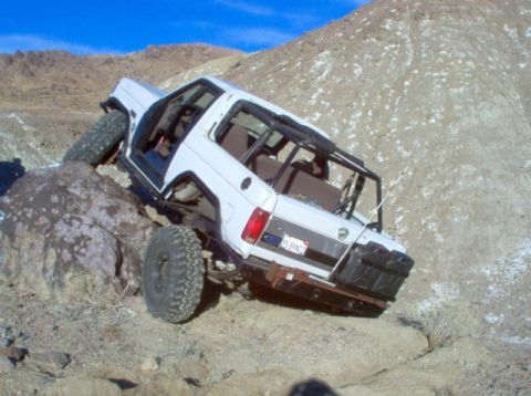 1988 Ford Bronco - This white SUV, identified by its license plate as a "95 BRONCO," navigates a challenging rocky terrain under a clear blue sky.