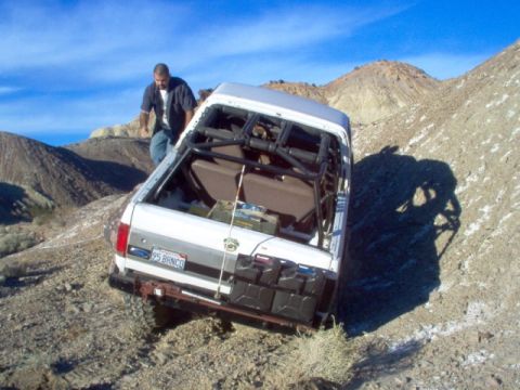 1988 Ford Bronco - A white Ford Bronco with a roll cage and multiple jerry cans is seen navigating a steep, rocky terrain.