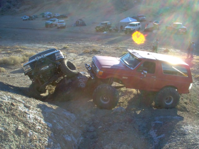 Red Ford Bronco and black truck climbing rocky terrain with tents and other vehicles in background