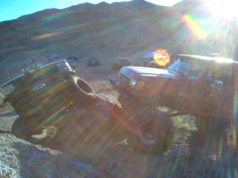 1988 Ford Bronco - Two off-road trucks are parked on a rocky terrain under a bright sun, with hills in the distance.