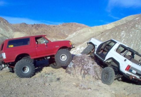 1988 Ford Bronco - Two modified off-road vehicles navigate a challenging rocky terrain under a clear blue sky.