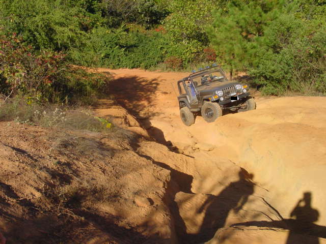 Gray Jeep Wrangler crawling over an orange sandstone ledge on a wooded trail, shadow of photographer