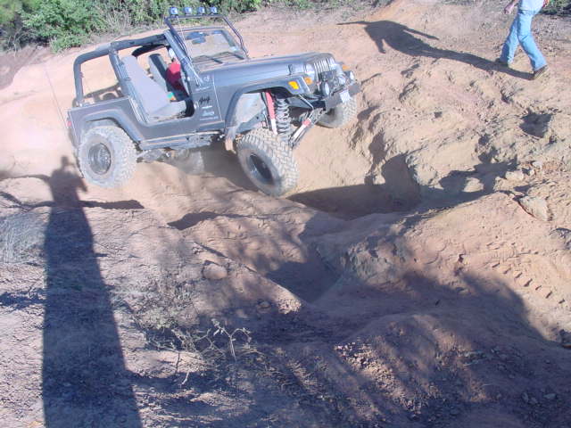 Doorless dark gray Jeep TJ on big off-road tires climbing down into a red dirt trench on a trail
