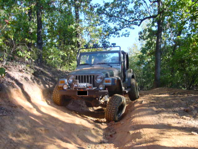 Camo-painted Jeep Wrangler with light bar climbing a dirt trail between trees
