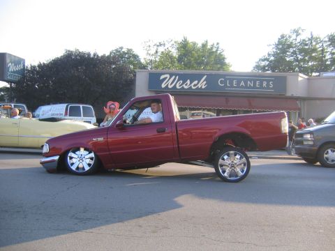 1978 Ford Bronco - A heavily modified red pickup truck displays its custom wheels and lowered stance in front of a business.