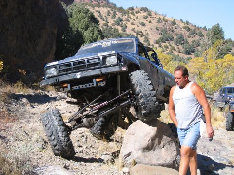 1986 Ford Bronco - A heavily modified off-road truck navigates a rocky trail, with a man walking nearby.