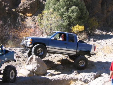 1986 Ford Bronco - A lifted blue pickup truck is shown off-roading on a rocky trail with sparse vegetation.