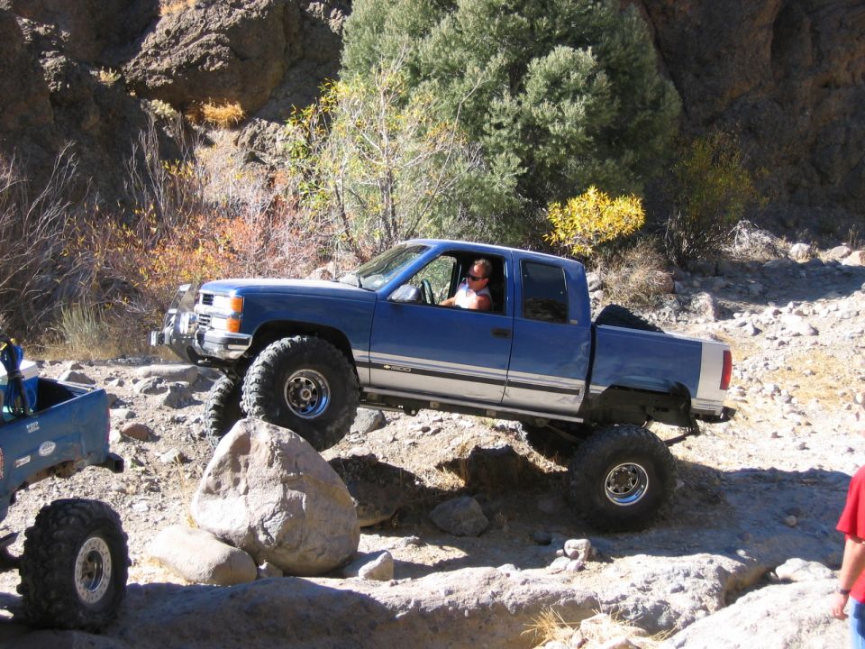 Blue pickup truck with large tires navigating rocky terrain. Another off-road vehicle is partially visible to the left