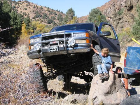 1986 Ford Bronco - A blue pickup truck is shown off-roading with its front end lifted over rocks.