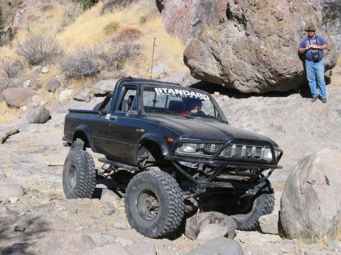 1986 Ford Bronco - A modified black pickup truck navigates a challenging rocky trail with a person observing in the background.