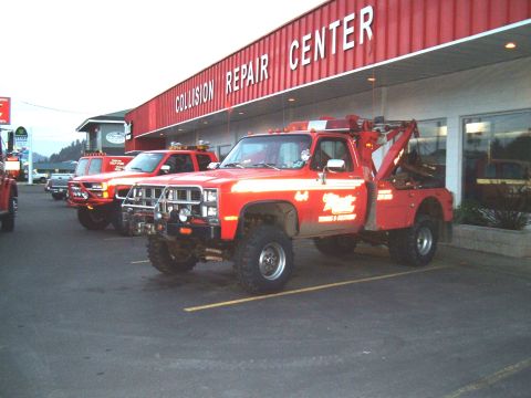 2000 Ford F-250 - A red tow truck with white stripes and a logo on its door is parked outside a collision repair center, with other trucks visible behind it.