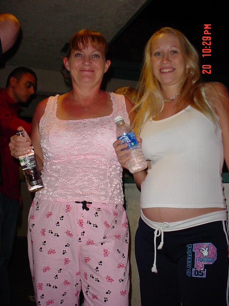 two women, one in pink pajamas, the other in a white tank top, holding drinks