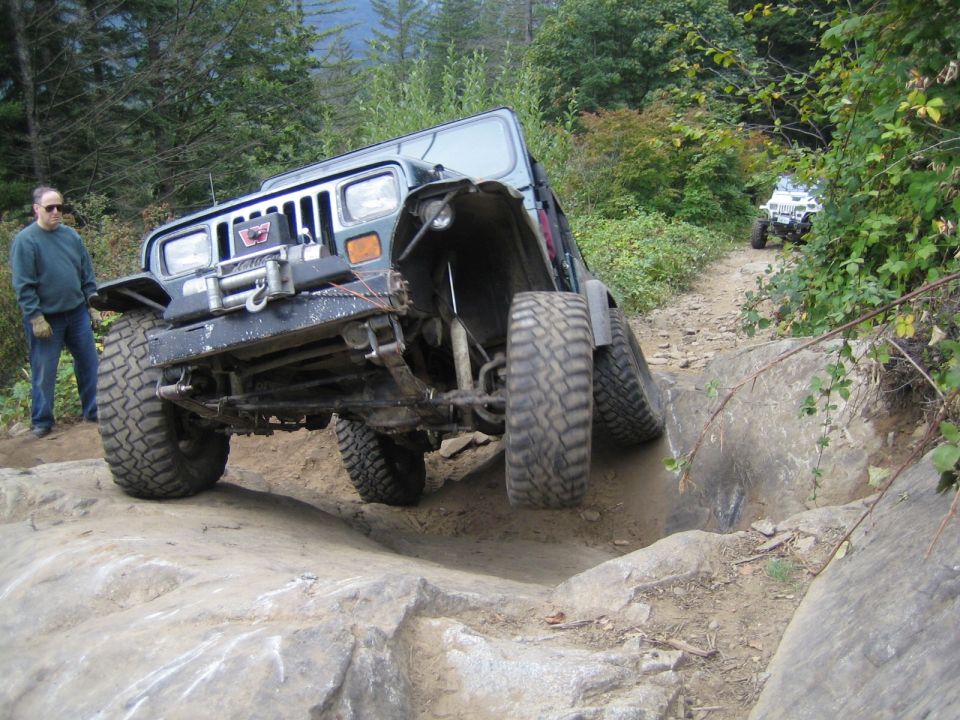 Jeep Wrangler navigating a difficult rocky trail with a person standing nearby; another ATV is visible further down the path