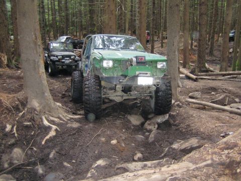 1990 Ford Bronco - Several off-road vehicles traverse a challenging, muddy path through a dense forest.