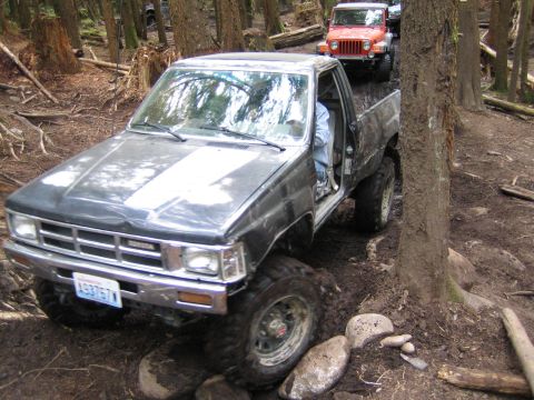 1990 Ford Bronco - Modified Toyota pickup truck with large tires navigating a muddy forest trail, another Jeep visible behind it.